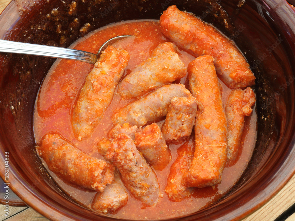 A brown bowl on a wooden table with a spoon containing a homemade sausages stew in spices and tomato sauce to eat with polenta, a typical Italian dish A brown bowl on a wooden table with a spoon containing a homemade sausages stew in spices and tomato sauce to eat with polenta, a typical Italian dish