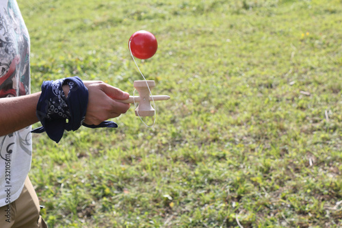 A young girl with a blue colored t-shirt holding a wooden kendama stick and playing with a red ball in a green grass lawn during a sunny summer