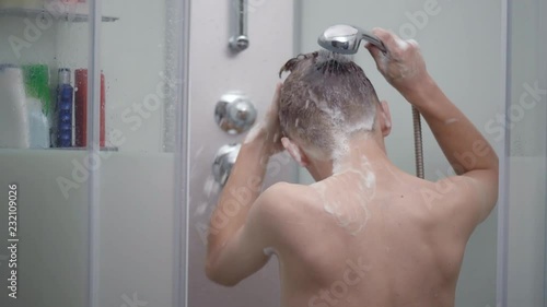 Young boy bathing under a shower at home - back view. Beautiful teen boy taking shower and washing in the bathroom. Happy child washing head, face and body with water.