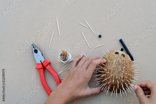 Hedgehog artwork DIY craft from coconut shell and toothpicks. Pliers, coconut shell, one black crayon and two hands with brown background.  Cool  art idea and activity for kids, teens and boys.
