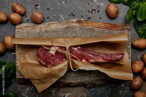 raw rib eye steak wrapped in baking paper on wooden board with spices and potatoes