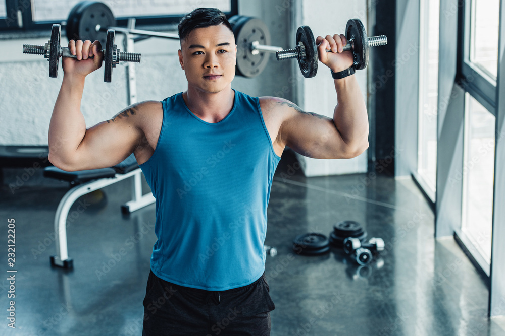 concentrated young sportsman exercising with dumbbells in gym and looking at camera concentrated young sportsman exercising with dumbbells in gym and looking at camera