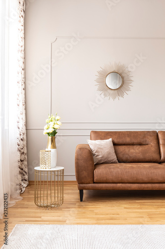 End table with fresh white roses and glass vase sanding by the window with curtains in real photo of bright sitting room interior with round mirror on the wall and brown leather sofa with cushion