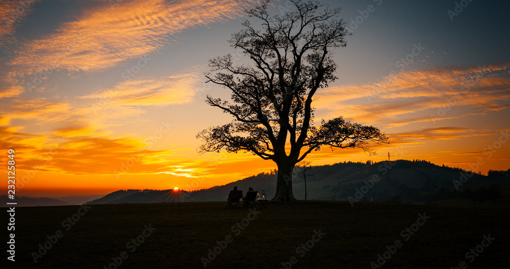 Loving couple watching sunset Great Lhota under an old lonely tree on a hill beautiful orange sunset sky full of clouds...