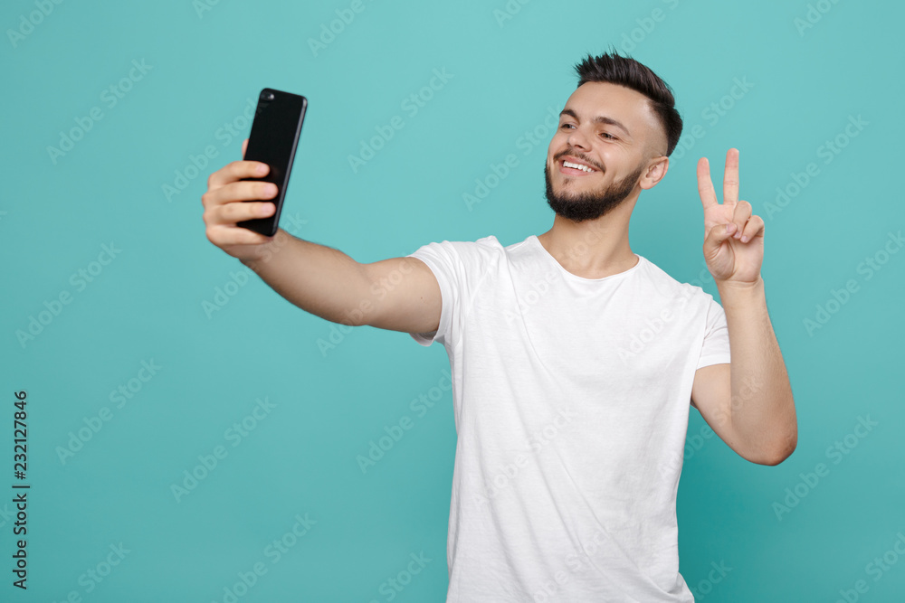 Handsome young bearded hipster in white t-shirt using phone and taking selfie with bright smile. Handsome young bearded hipster in white t-shirt using phone and taking selfie with bright smile.