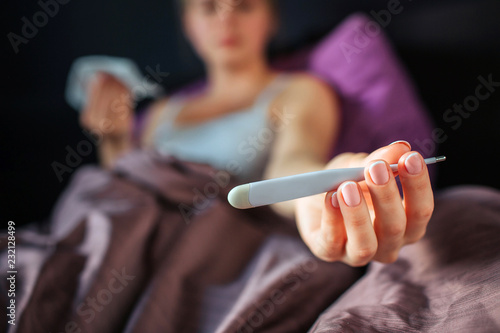 Young woman has temperature. She holds thermometer in hand. Blonde holds white tissue in another hand. She looks at thermometer. Model stays in bed.