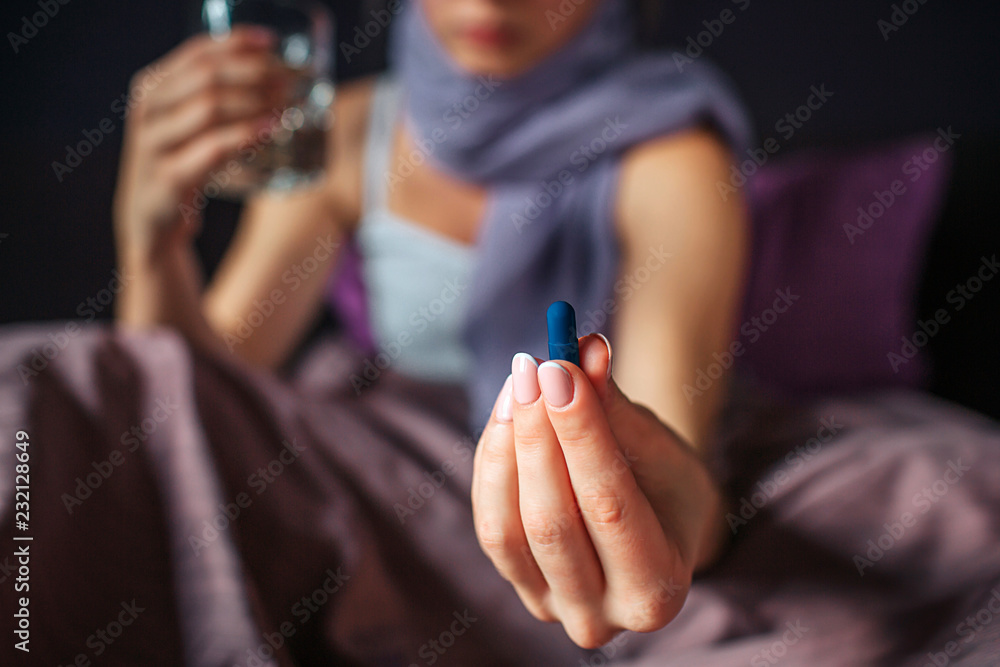 Obraz premium Cut view of young woman sits on bed and shows medicine in hand. It is blue capsule. There is glass of water in another hand. Her neck is covered with scarf.