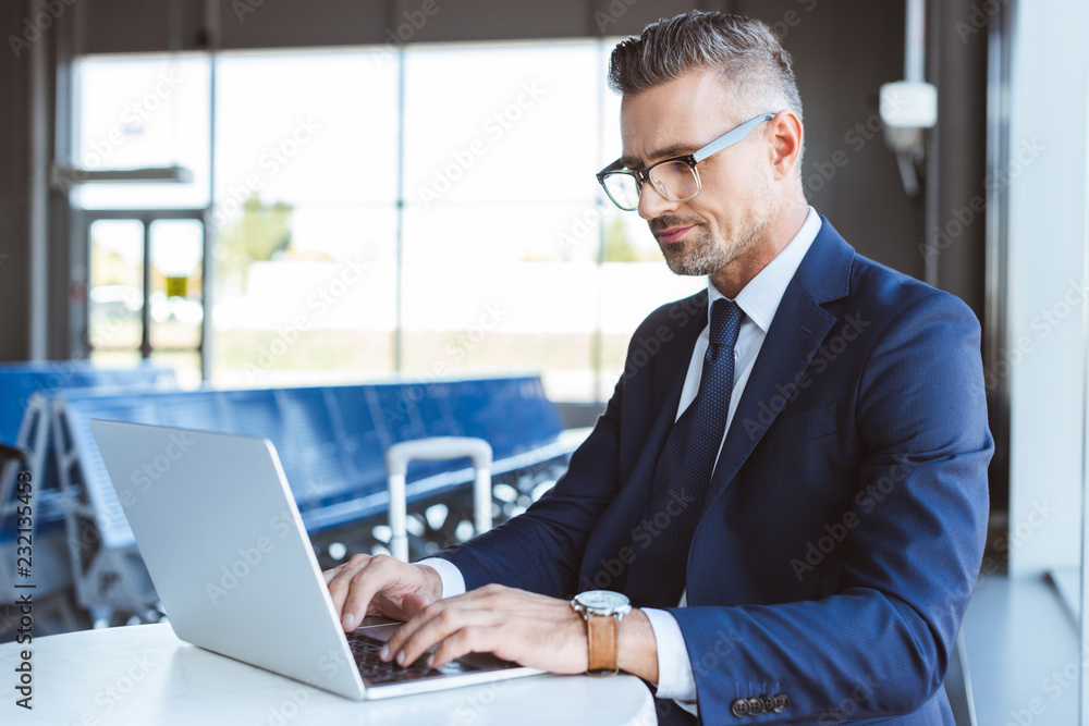 handsome businessman in glasses typing on laptop in airport handsome businessman in glasses typing on laptop in airport