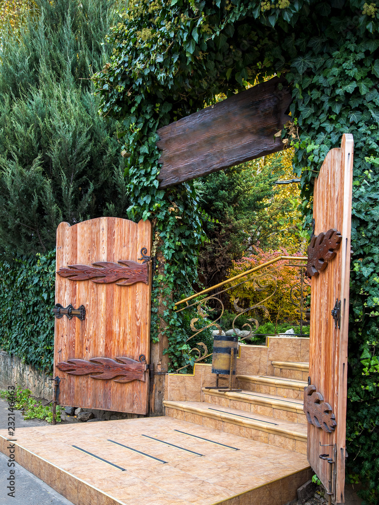 Naklejka premium The entrance to the wooden gate is overgrown with ivy and other evergreens, the passage to the territory of the house is tiled in front of the steps.