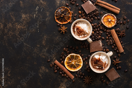 Two cups of hot chocolate on a dark background with roasted coffee beans, cinnamon sticks and sliced orange. Top view, copy space.