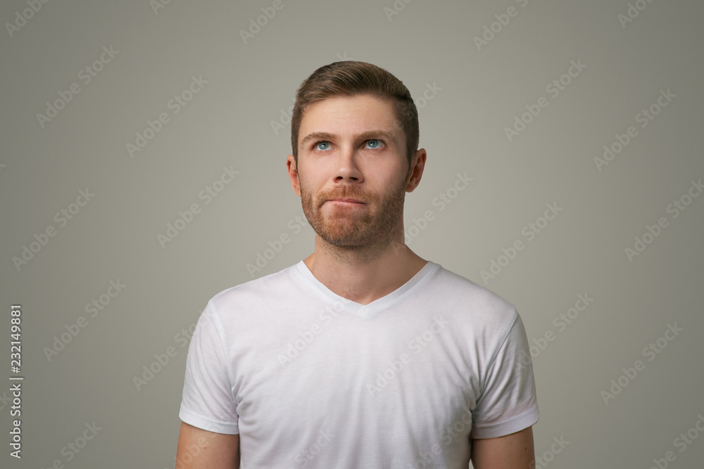 Studio portrait of a thoughtful young man in a white t-shirt. He presses his lips together hard and looks up. Isolated large image on grey background Studio portrait of a thoughtful young man in a white t-shirt. He presses his lips together hard and looks up. Isolated large image on grey background