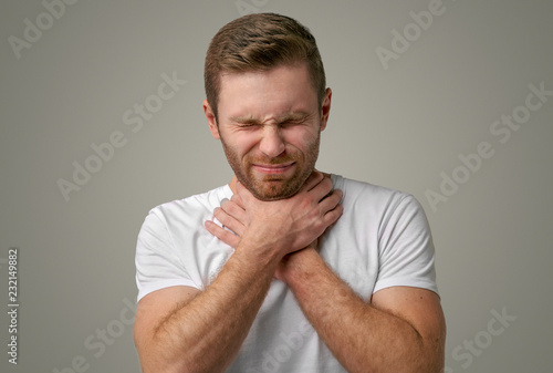 A man with a beard grabs his throat with both hands and squints intensely from the pain in his throat. The concept of depression, sore throat, choking and suicide