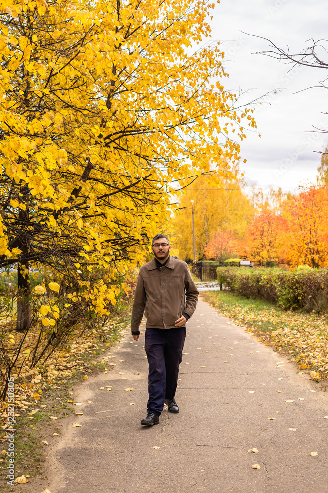 A young man with a beard and a mustache with glasses in a brown jacket, walking along the path in the autumn Park