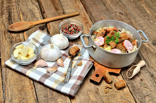 Close-up of garlic soup in an old saucepan, fresh garlic bulbs, cloves, colored pepper, wooden spoon, croutons and a towel on a rustic wooden table photo