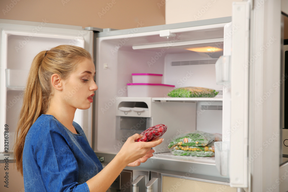 Young woman with frozen cherries near open refrigerator at home