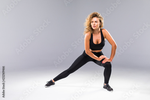 Portrait of sporty athletic woman in sneakers and tracksuit squatting doing sit-ups in gym isolated over gray background