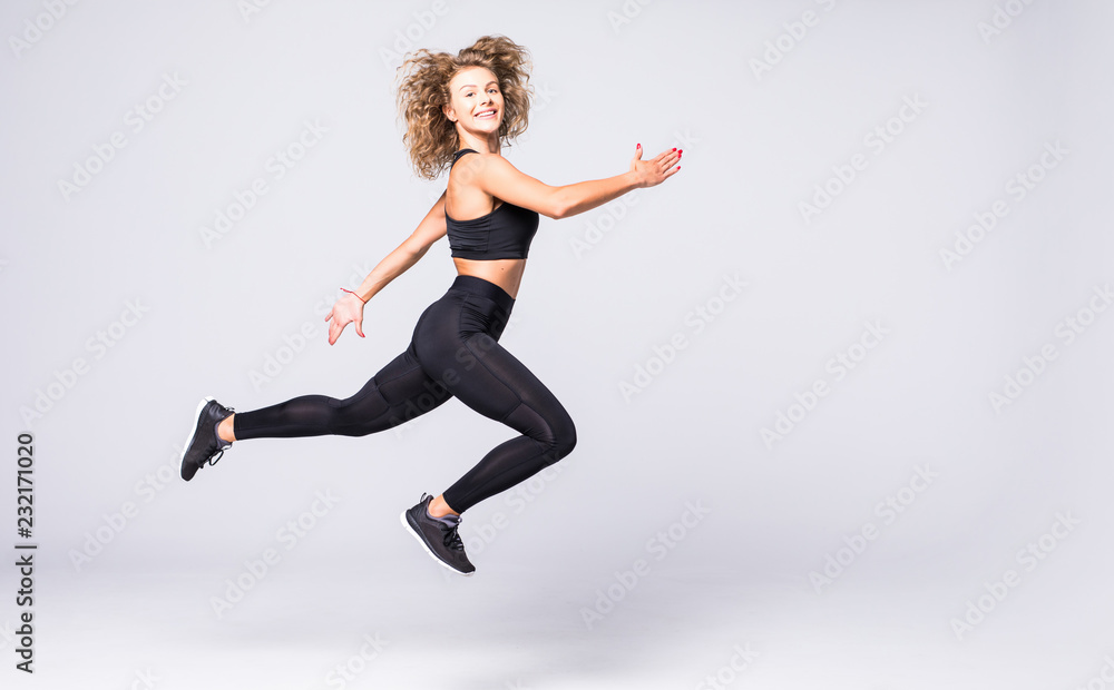 Beautiful young fit woman with curly hair, happily jumping on a grey background. Slim woman in sport clothes.