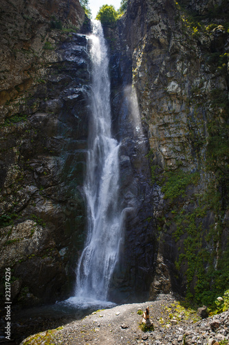 A high waterfall with white splashes of water descends from the mountain and sparkles in the sun. Attraction of Georgia Gveleti waterfall