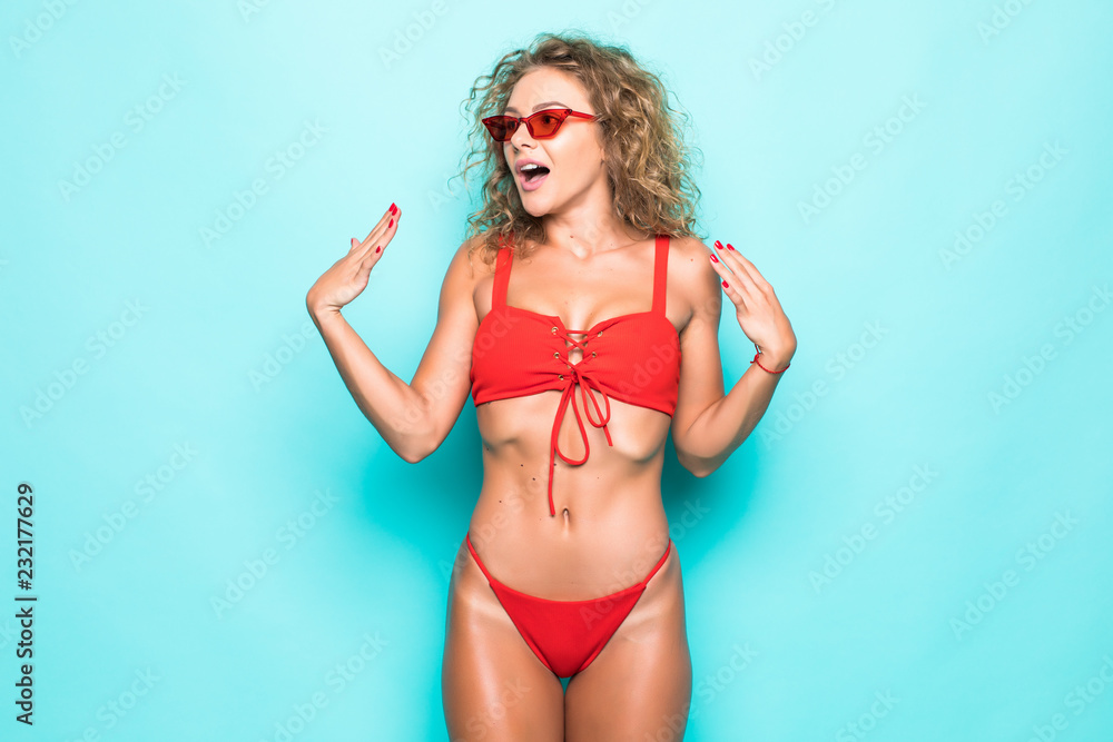 Beautiful surprised curly girl in colorful bikini and sunglasses, emotionally looking on camera over blue background Beautiful surprised curly girl in colorful bikini and sunglasses, emotionally looking on camera over blue background
