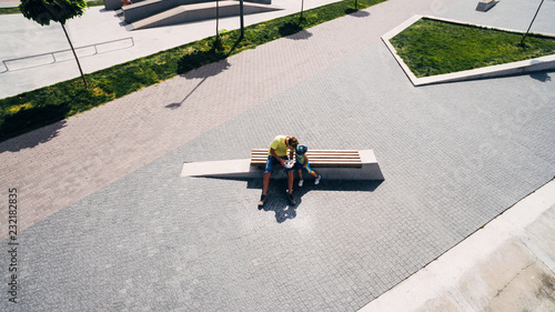 Aerial view. The father shows the child how he controls the drones. A father and a little son are sitting on a bench near a lake on a city beach