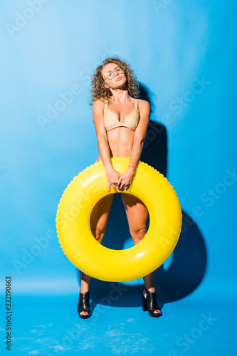 Young sexy woman with perfect body in bikini with yellow inflatable ring isolated over blue background