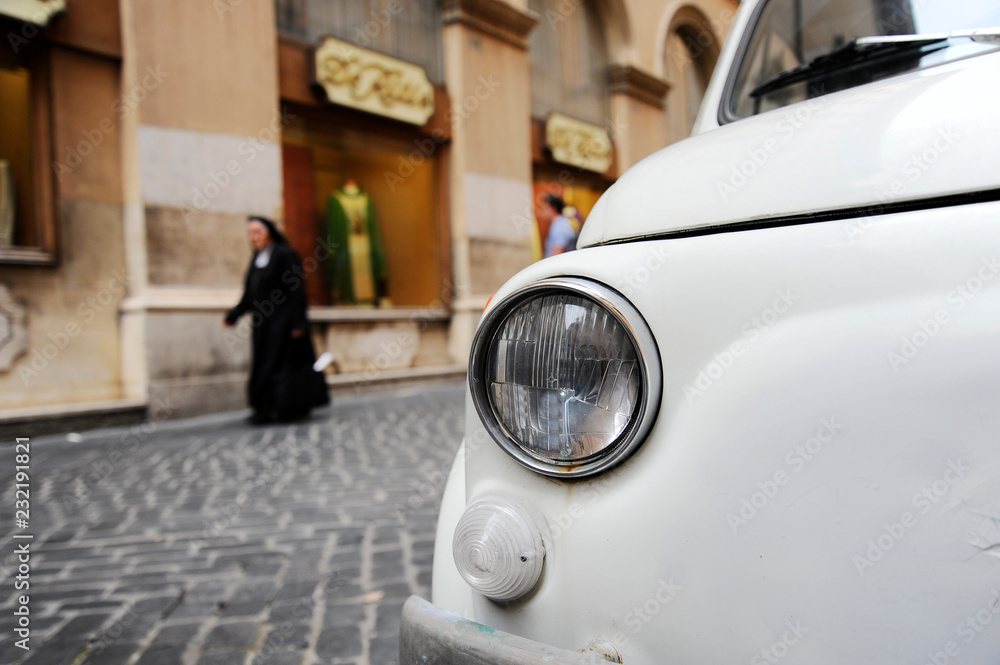 A parked vintage car in the streets of Rome, a nun walking on the background. The Fiat 500 Cinquecento is a famous Italian symbol.