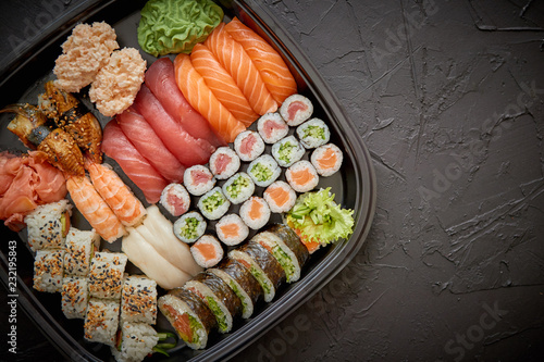 Japanese food concept. Catering, various kinds of sushi on plate or platter set. Chopsticks, ginger, soy sauce, wasabi. Placed on stone black background in plastic delivery box.