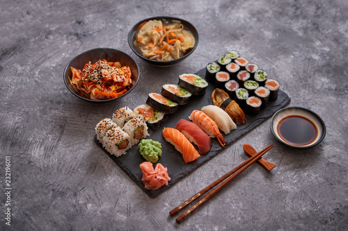 Japanese food composition. Various kinds of sushi placed on black stone board. Spicy kimchi salad, wontong soup, chopsticks and soy souce bowl.