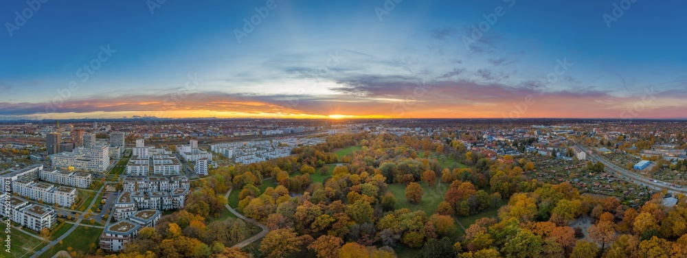 Fototapeta premium Urbanes Panorama eines Sonneruntergangs über der bayrischen Landeshauptstadt München im bunt beblätterten Hirschgarten, ein Park mit angrenzenden Wohnhäusern als Luftbild einer Drohne im Herbst