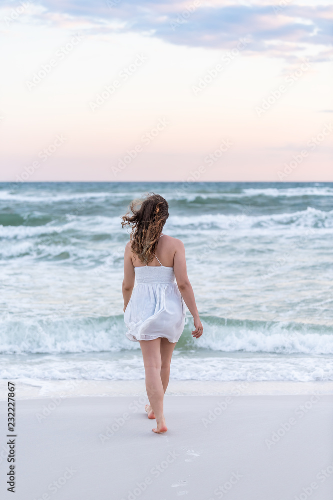 Young woman in white dress on beach pink sunset in Florida panhandle with wind, ocean waves, walking legs back to water Young woman in white dress on beach pink sunset in Florida panhandle with wind, ocean waves, walking legs back to water