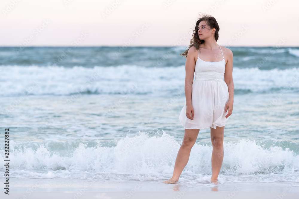 Young woman front standing in white dress on beach sunset in Florida panhandle with wind, ocean waves crashing legs, tan skin, looking to side sad Young woman front standing in white dress on beach sunset in Florida panhandle with wind, ocean waves crashing legs, tan skin, looking to side sad