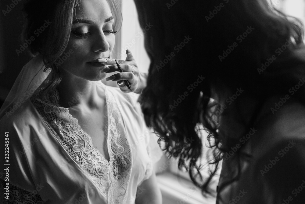 Stylish bride getting her make-up in the morning at window light. Makeup artist painting lips with brush in nude lipstick on bride face close-up. Stylish bride getting her make-up in the morning at window light. Makeup artist painting lips with brush in nude lipstick on bride face close-up.
