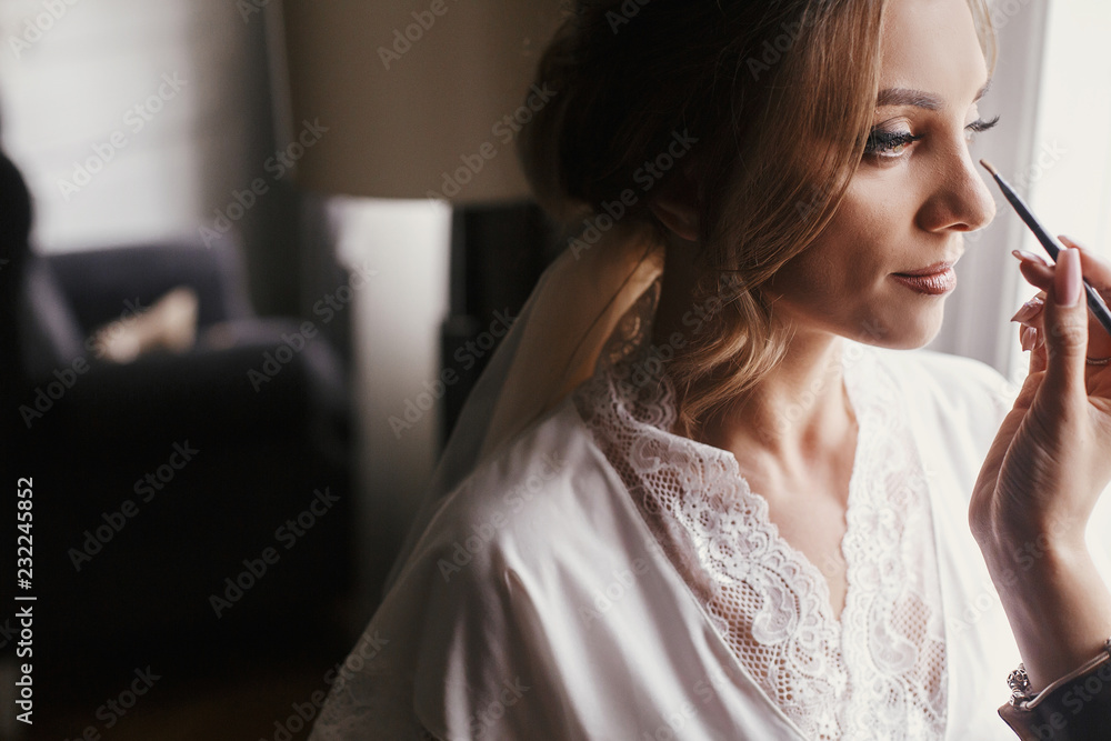 Stylish bride getting her make-up in the morning at window light. Makeup artist doing brows and applying eyeshadow on bride face close-up. Stylish bride getting her make-up in the morning at window light. Makeup artist doing brows and applying eyeshadow on bride face close-up.