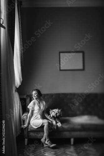 Gorgeous beautiful bride in silk robe holding modern bouquet and sitting on stylish gray sofa. Happy stylish Bride with perfect makeup posing near window in soft light in the morning