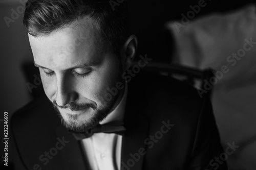 Stylish groom in black suit and bow tie, sitting in chair at window light. Confident and happy portrait of man. Groom getting ready in morning