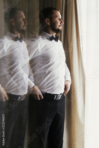 Stylish groom in white shirt and bow tie posing at window light. Confident and happy portrait of man. Groom getting ready in morning. Creative wedding photo with mirror reflection