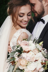 Gorgeous bride and stylish groom gently hugging and smiling on background of green trees. Sensual wedding couple embracing. Romantic moments of newlyweds. Wedding photo