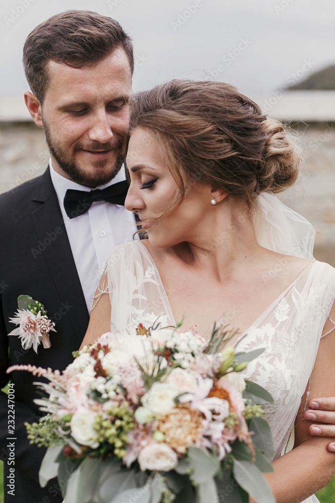 Gorgeous bride with modern bouquet and stylish groom gently hugging and smiling outdoors. Sensual wedding couple embracing. Romantic moments of newlyweds. Wedding photo Gorgeous bride with modern bouquet and stylish groom gently hugging and smiling outdoors. Sensual wedding couple embracing. Romantic moments of newlyweds. Wedding photo