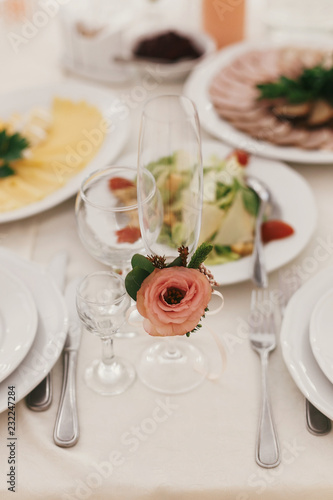 Stylish empty glasses and plate, cutlery,and floral decor on table at wedding reception in restaurant. Luxury wedding catering. Modern wedding setting