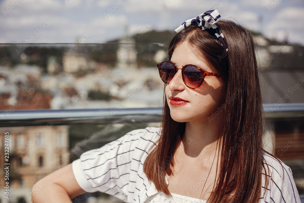 Beautiful stylish young woman in  sunglasses, retro dress and headband relaxing on roof in european city street. Hipster girl smiling and enjoying summer time in Lviv. Space for text