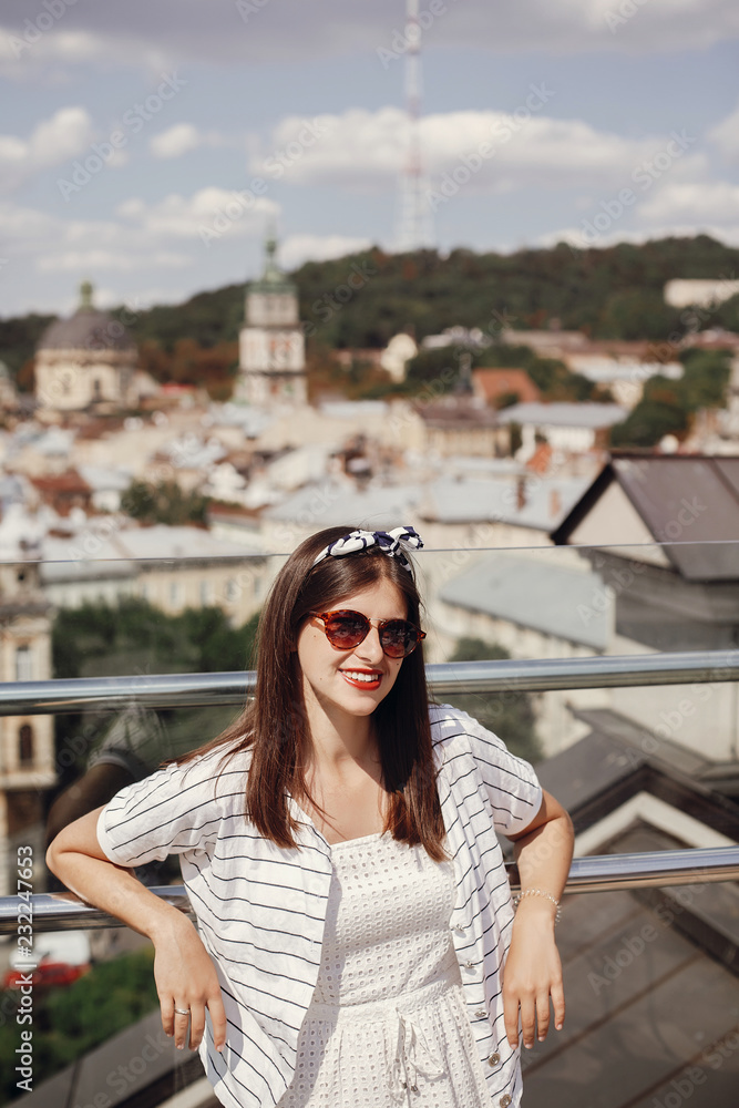 Beautiful stylish young woman in sunglasses, retro dress and headband relaxing on roof in european city street. Happy hipster girl smiling and enjoying summer time in Lviv. Copy space