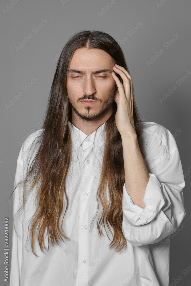 Vertical portrait of creative young male painter or musician with beard and long loose hair closing eyes and touching his head while having bad terrible headache because of stress or problem Vertical portrait of creative young male painter or musician with beard and long loose hair closing eyes and touching his head while having bad terrible headache because of stress or problem
