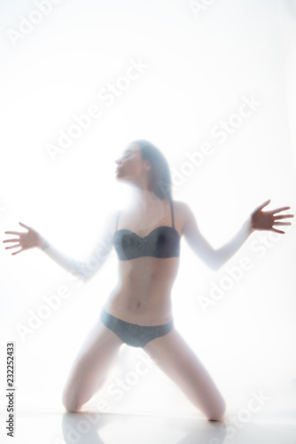 Young beautiful woman posing in lingerie behind translucent material. Full-length attractive sexy girl's portrait in studio over white background, foggy silhouette. Art female portrait.
