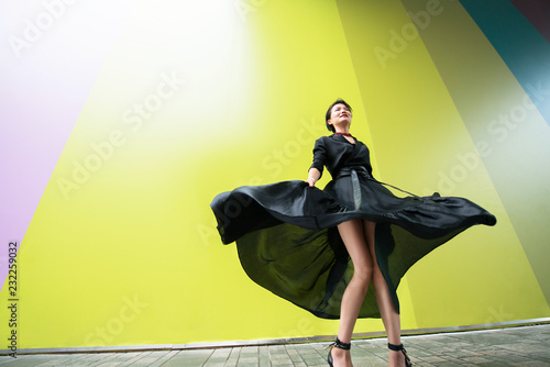 Fashion woman. Young beautiful chinese girl dancing outdoor wearing long black dress with high heels over colorful wall background. Stylish trendy lady. photo