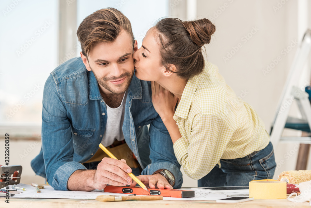 Fototapeta premium young woman kissing smiling man marking blueprint with level tool