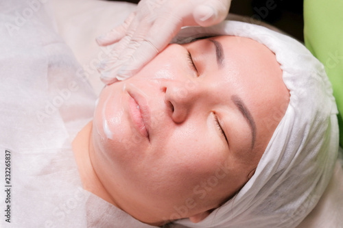 Cosmetician's hand smears the cream on the cheek of an Asian girl. Eastern woman lies on the procedure of rejuvenation of the skin in the beauty salon.