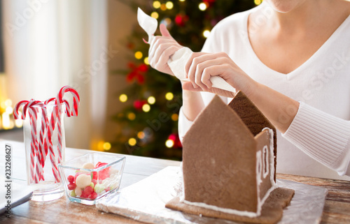 cooking, holidays and people concept - woman with pastry bag making gingerbread houses at home over christmas tree lights background