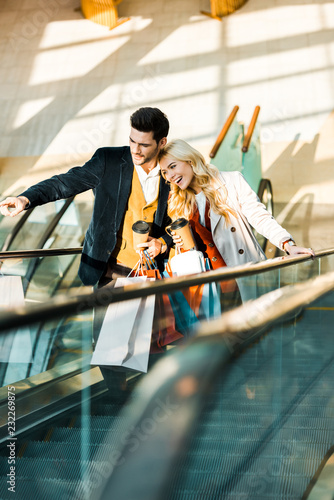 happy stylish man with coffee to go showing something to girlfriend with shopping bags standing on escalator