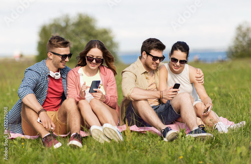 friendship, technology and leisure concept - group of smiling friends with smartphones sitting on grass in summer