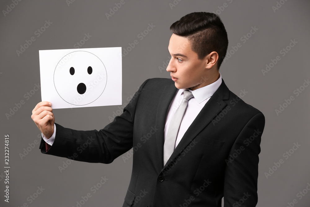 Businessman holding sheet of paper with drawn emoticon on grey background Businessman holding sheet of paper with drawn emoticon on grey background